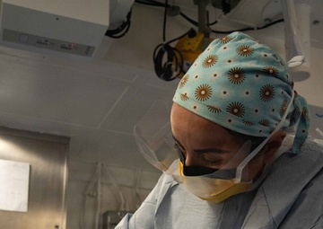 A medical professional, likely a scrub technician or surgical nurse, is working at a sterile instrument table in what appears to be an operating room. They are wearing full surgical PPE including a patterned scrub cap, face shield, surgical mask, gown, and double-gloved hands. The individual appears to be organizing or preparing surgical instruments and labeling items prior to or during a surgical procedure.