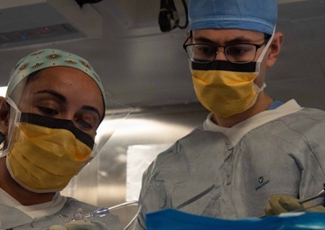 Two medical professionals in surgical attire are performing or assisting with a medical procedure in what appears to be an operating room or clinical setting. Both individuals are wearing yellow surgical masks with black eye shields, blue/gray surgical gowns branded 'HALYARD', and latex gloves. One person is handling clear tubing while the other is working with a surgical instrument near a blue sterile drape.