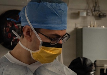 A medical professional wearing full sterile attire is handling what appears to be a thin wire or catheter connected to a small device or syringe. The individual is dressed in a blue surgical cap, yellow face mask, glasses, a white disposable gown, and yellow latex gloves. The setting appears to be a clinical or surgical environment with medical equipment visible in the background.