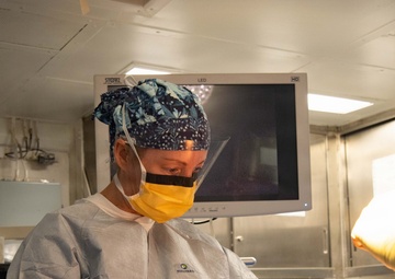 A medical professional in full surgical attire is performing a surgical procedure in an operating room. They are wearing a Halyard-branded surgical gown, yellow face mask, face shield, surgical cap, and latex gloves while working on a patient covered with blue surgical draping. A Storz LED HD monitor is visible in the background.