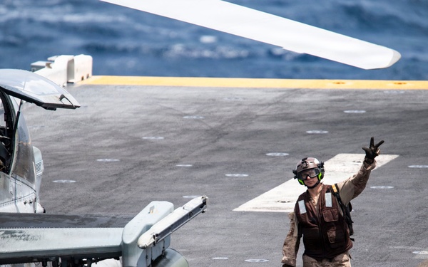 A military deck crew member is signaling with a raised hand on what appears to be the flight deck of a naval vessel, likely an amphibious assault ship. In the foreground, a chained-down attack helicopter — identifiable as a Bell AH-1Z Viper — is visible with weapons systems including rocket pods and missiles mounted on its stub wings. The ocean is visible in the background, confirming an at-sea operational setting.