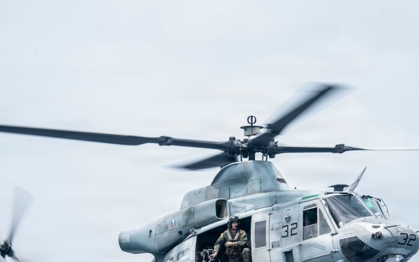 A U.S. Marines UH-1Y Venom helicopter (tail number 32) is captured in flight against an overcast sky. The aircraft belongs to VMM-163 squadron and has its side door open with a crew member/gunner seated at the door position. The helicopter is in a gray low-visibility paint scheme typical of modern Marine Corps aviation.