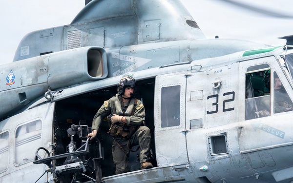 A military helicopter, numbered '32', is captured in flight with an open side door. A crew member in flight gear and helmet is standing in the doorway, operating or monitoring what appears to be a mounted camera or sensor system. The aircraft displays a 'RESCUE' marking and a unit insignia patch on its fuselage.