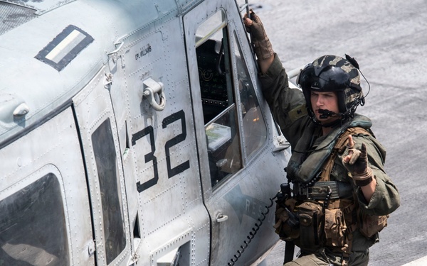 A military personnel member in full tactical gear is standing beside a military helicopter marked with the number '32' on what appears to be a ship's flight deck. The individual is wearing a flight helmet with communications equipment and is gesturing with one hand while gripping the helicopter frame with the other. The scene suggests a pre-mission briefing, boarding preparation, or coordination activity.