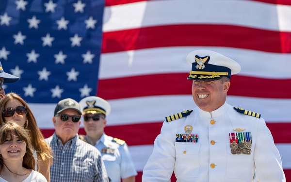 A senior Coast Guard officer in full dress white uniform walks past seated and standing guests at what appears to be a formal military ceremony. A large American flag serves as a backdrop, and a formation of uniformed personnel is visible on the right side. The officer is smiling and interacting with civilian attendees, including an elderly man and a young girl.