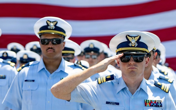 A formation of United States Coast Guard personnel in tropical blue uniform (short-sleeve light blue shirts with navy trousers) stand at attention during a formal ceremony. The foreground officer, wearing a commander or lieutenant commander rank insignia and service ribbons, is rendering a salute. A large American flag is prominently displayed in the background.