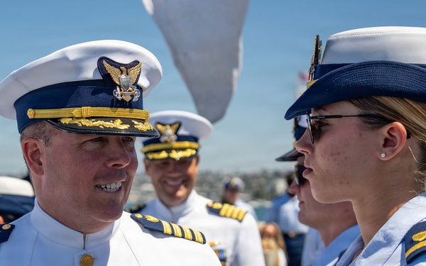 A formal military ceremony featuring United States Coast Guard personnel in dress uniforms. A senior officer in white dress uniform with multiple medals and ribbons is engaged in conversation with a junior officer in light blue service uniform. The setting appears to be an outdoor waterfront location on a clear, sunny day.