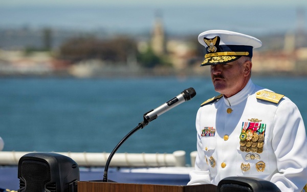 A high-ranking U.S. Coast Guard officer is delivering a speech at a podium aboard the USCGC Munro (WMSL 755), a Legend-class national security cutter. The ceremony appears to be a formal military event, likely a change of command or commissioning ceremony, taking place on the ship's deck with a coastal cityscape visible in the background. A second uniformed officer is seated behind the speaker.