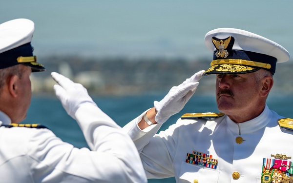 Two senior military officers in white dress uniforms are exchanging salutes in a formal ceremony, likely aboard a vessel or waterfront location. The officer facing the camera displays an extensive array of medals, ribbons, and qualification badges indicating high rank and long service. The setting appears to be an outdoor change-of-command or retirement ceremony based on the formal attire and mutual salute.