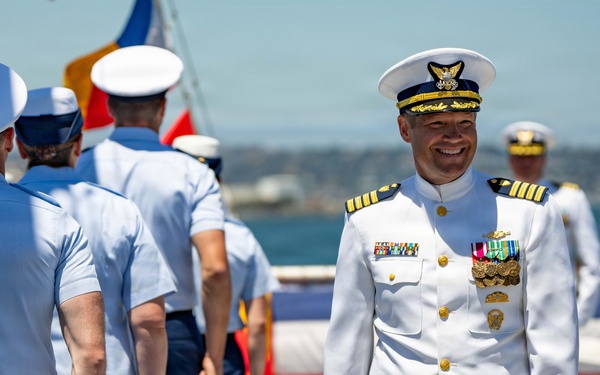 A formal military ceremony is taking place on what appears to be the deck of a naval vessel near a waterfront. A senior officer in a white dress uniform with gold insignia, medals, and a ceremonial sword stands prominently smiling, while enlisted personnel in light blue shirts and navy trousers stand in formation. The setting suggests a change of command, commissioning, or similar official military ceremony.