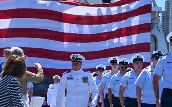 A formal U.S. Coast Guard ceremony is taking place on what appears to be the deck of a vessel or pier, with a large American flag displayed prominently in the background. A senior officer in dress whites with gold shoulder boards and service ribbons walks down a central aisle flanked by uniformed personnel standing at attention. Civilian guests are seated or standing on the left side, suggesting this is a change of command, commissioning, or similar official military ceremony.