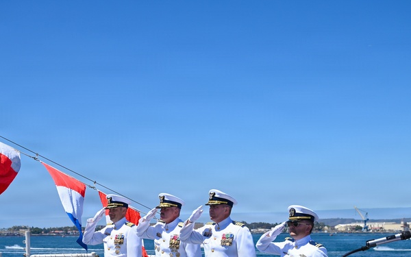 A formal U.S. Navy change of command or commissioning ceremony is taking place on the deck of a naval vessel, likely in San Diego harbor based on the visible waterfront and naval facilities in the background. Four naval officers in white dress uniforms are rendering salutes while a woman in a red top stands at a podium, likely performing the national anthem or reading orders. Signal flags are strung across the ship's deck as decoration for the ceremony.