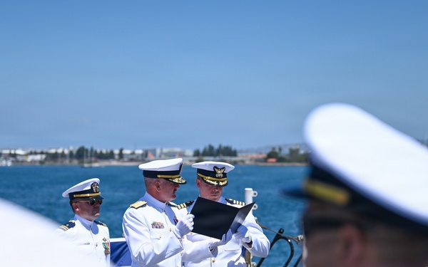 A formal military ceremony is taking place on the deck of a naval vessel, likely a change of command or commissioning ceremony. Two officers in white dress uniforms are reading from a black folder, while other uniformed personnel stand at attention. The setting appears to be a harbor or bay with a clear blue sky and waterfront visible in the background.