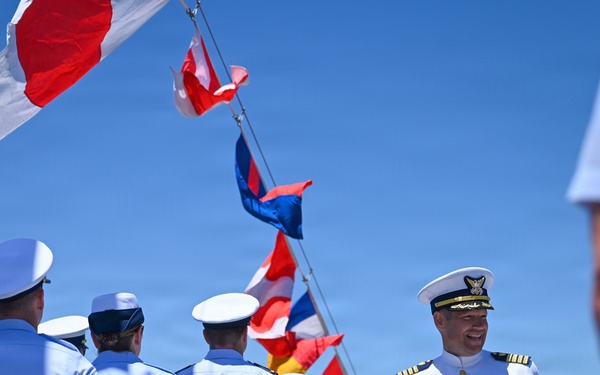 A formal military ceremony is taking place outdoors on what appears to be the deck of a vessel or waterfront location on a clear, sunny day. A senior officer in a white dress uniform with gold insignia and multiple medals is prominently featured, smiling while facing a formation of personnel in light blue shirts and dark trousers. Multiple nautical signal flags are displayed in the background, suggesting a commissioning, change of command, or similar naval/Coast Guard ceremony.