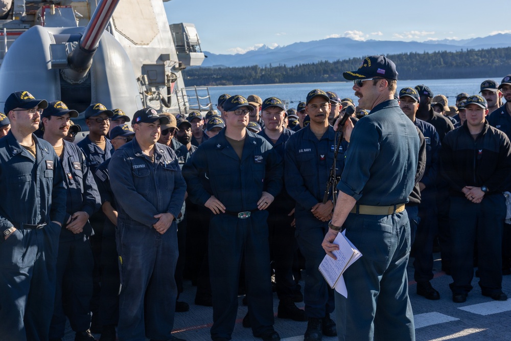 A U.S. Navy officer addresses a large group of sailors assembled on the deck of a naval vessel, likely a destroyer or guided-missile cruiser, identifiable by the visible gun turret. The gathering appears to be an all-hands or muster formation, with the officer holding papers and speaking into a microphone. The setting appears to be a Pacific Northwest naval installation, with forested shoreline and snow-capped mountains visible in the background.
