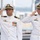 Two U.S. Navy officers in white dress uniforms are rendering a salute simultaneously, likely during a formal military ceremony. Both wear officer cover caps with gold insignia and display multiple medals, ribbons, and qualification badges on their uniforms. The background suggests an outdoor naval or waterfront setting.