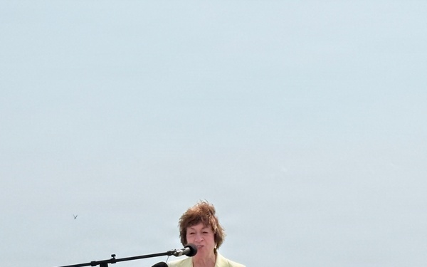 A woman in a yellow blazer stands at a wooden podium giving a speech or press conference outdoors near a rocky coastline. Multiple microphones are mounted on the podium and a boom microphone extends from a stand to her left. The setting appears to be a sunny day with calm ocean water visible in the background.