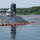 A U.S. Navy submarine, identified as USS South Dakota (SSN-790), nicknamed 'The Gunslingers,' is moored at a pier in calm waters surrounded by a bright orange containment/oil boom. Personnel and a white tent canopy are visible on the deck near the sail (conning tower). The setting appears to be a naval facility with dense green tree lines in the background.