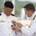 Two naval officers in white dress uniforms are engaged in a medal pinning ceremony. One officer is attaching or adjusting a medal and ribbon onto the chest of the other officer, who is looking down at the decoration being applied. The setting appears to be an outdoor military ceremony with a waterfront or harbor visible in the background.