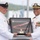Two U.S. Navy personnel in white dress uniforms are participating in a formal military ceremony, jointly holding a shadow box containing a folded flag and a commemorative plaque. The officer on the right displays multiple medals and ribbons, a command insignia, and shoulder boards indicating senior rank (Captain), while the individual on the left wears a Chief Petty Officer combination cover. The setting appears to be outdoors near a waterfront, consistent with a change of command or retirement ceremony.