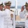 Three U.S. Navy officers in white dress uniforms are participating in a formal military ceremony, likely a change of command or commissioning event. Two officers are exchanging salutes while a third stands at attention between them. The setting appears to be an outdoor naval facility or ship deck.
