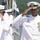 A senior U.S. Navy officer in white dress uniform is rendering a salute during what appears to be a formal military ceremony. He is adorned with numerous medals, ribbons, and insignia indicating high rank, likely a flag officer (Admiral). Two other naval personnel in white uniforms and covers are also saluting in the foreground.