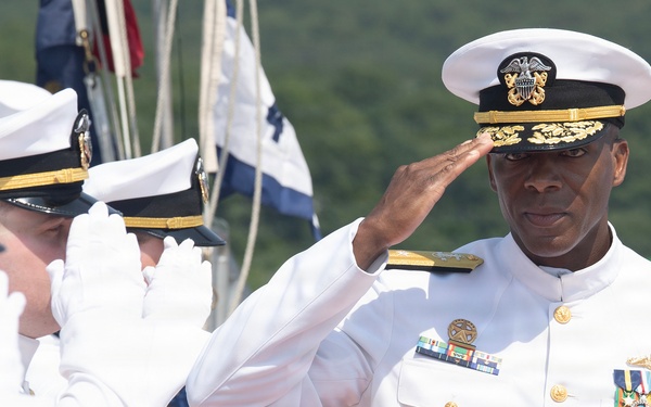 A senior U.S. Navy officer in white dress uniform is rendering a salute during what appears to be a formal military ceremony. He is adorned with numerous medals, ribbons, and insignia indicating high rank, likely a flag officer (Admiral). Two other naval personnel in white uniforms and covers are also saluting in the foreground.