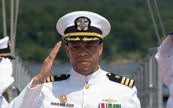 A U.S. Navy officer in white dress uniform is rendering a salute during what appears to be a formal military ceremony, likely a change of command or sideboy honors on a pier or dock. The central officer displays multiple medals and decorations along with gold shoulder boards indicating senior rank. Several other naval personnel in white dress uniforms flank him, also saluting.
