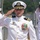A U.S. Navy officer in white dress uniform is rendering a formal salute during what appears to be a military ceremony. The central figure displays multiple medals, ribbons, and insignia consistent with a senior officer rank. Several other naval personnel in white dress uniforms are visible in the background, also saluting.