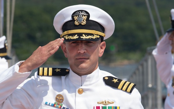 A U.S. Navy officer in white dress uniform is rendering a formal salute during what appears to be a military ceremony. The central figure displays multiple medals, ribbons, and insignia consistent with a senior officer rank. Several other naval personnel in white dress uniforms are visible in the background, also saluting.