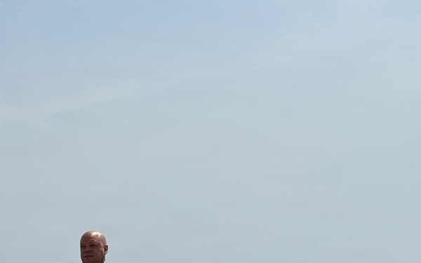 A bald man in a dark suit and tie is speaking at a wooden podium equipped with multiple microphones, positioned outdoors near a rocky shoreline. The backdrop features a large body of water, likely an ocean or large lake, with what appears to be a construction barge or vessel visible in the distance to the right. The setting suggests an official press conference or public announcement held at a waterfront location.
