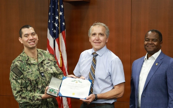 Three individuals are posed together in what appears to be an official military or government ceremony. A U.S. Navy servicemember in digital camouflage uniform (NWU Type III) stands alongside a civilian holding a certificate and medal case, with a third man in a business suit standing to the right. An American flag is visible in the background, suggesting a formal award or recognition presentation.