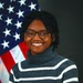 A formal portrait photograph of a smiling individual posed in front of a United States flag against a dark background. The subject is wearing a dark gray turtleneck sweater with white horizontal stripes and black-framed glasses. The composition and flag backdrop suggest this is an official or professional headshot.