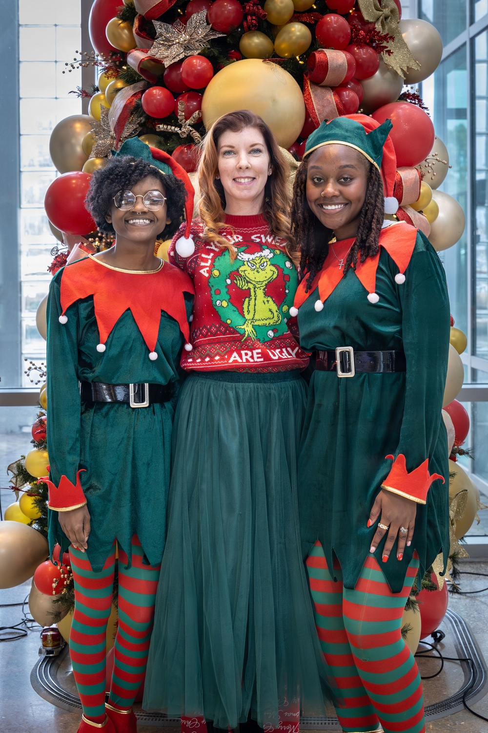 Three women pose together in front of a large Christmas balloon and ornament display. Two women on the left and right are dressed in matching green and red elf costumes with striped stockings and elf hats, while the woman in the center wears a Grinch-themed ugly Christmas sweater and a green tulle skirt. The setting appears to be an indoor venue with large windows visible in the background.