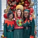 Three women pose together in front of a large Christmas balloon and ornament display. Two women on the left and right are dressed in matching green and red elf costumes with striped stockings and elf hats, while the woman in the center wears a Grinch-themed ugly Christmas sweater and a green tulle skirt. The setting appears to be an indoor venue with large windows visible in the background.