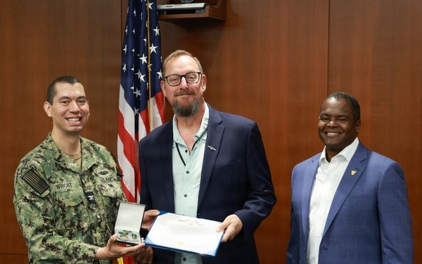 Three individuals are posing for a formal photograph in what appears to be an official government or military setting. A U.S. Navy sailor in digital camouflage uniform (NWU Type III) is presenting a medal in a display case and a certificate to a civilian man in a blue blazer, while a third man in a blue suit stands to the right. The American flag and wood-paneled walls suggest an official award or recognition ceremony.