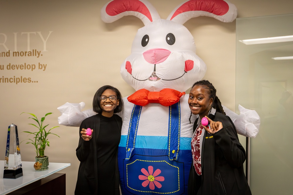 Two young women are posing and smiling alongside a large inflatable Easter bunny costume in what appears to be an office or institutional lobby. Each woman is holding a small pink Easter egg. The setting includes motivational text on the wall and a small plant on a reception desk.