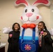 Two young women are posing and smiling alongside a large inflatable Easter bunny costume in what appears to be an office or institutional lobby. Each woman is holding a small pink Easter egg. The setting includes motivational text on the wall and a small plant on a reception desk.