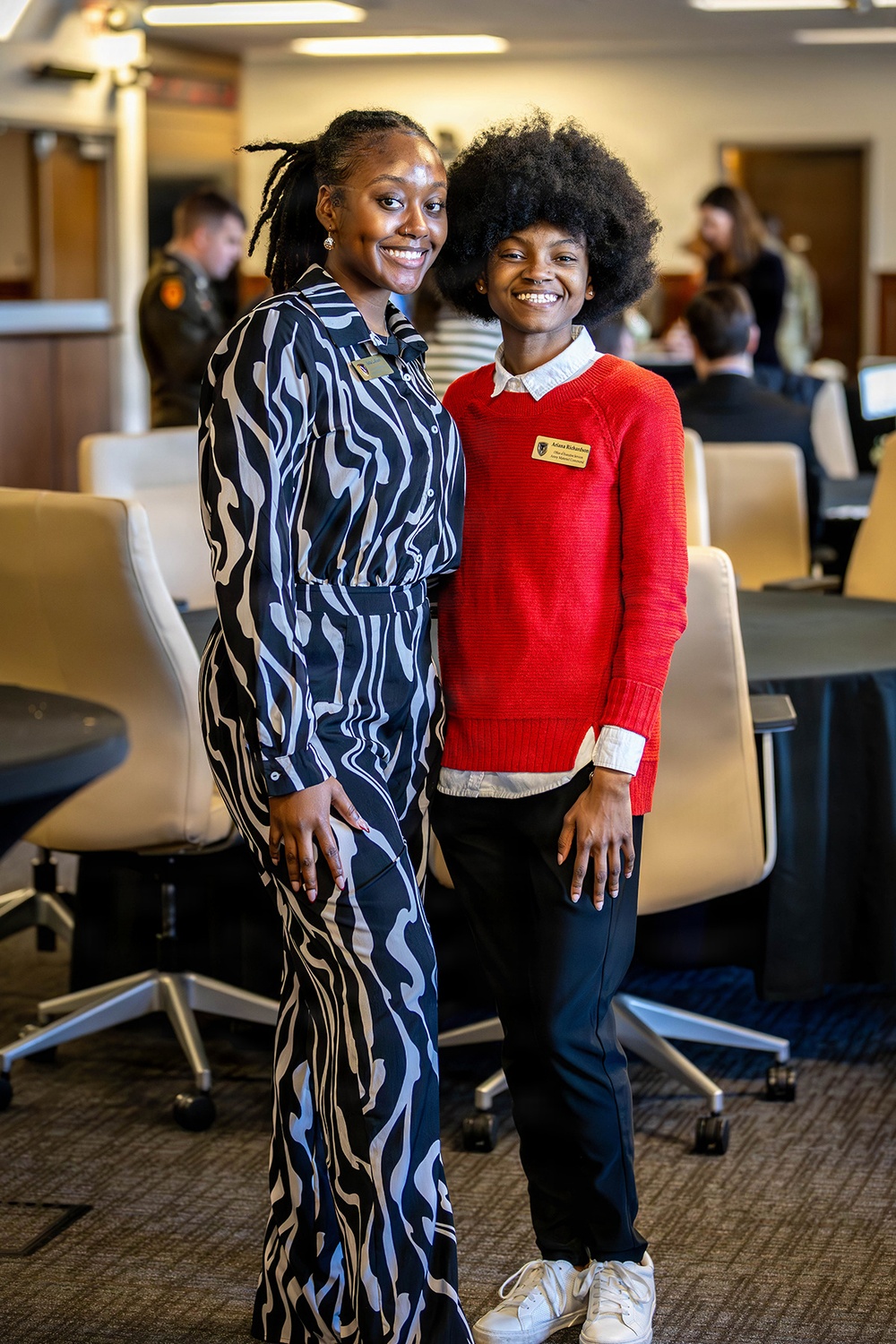 Two young women pose together smiling in what appears to be a professional or academic event setting. Both are wearing name badges, suggesting an organized gathering such as a conference, orientation, or networking event. The background shows additional attendees seated at tables in a well-lit room.