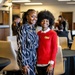 Two young women pose together smiling in what appears to be a professional or academic event setting. Both are wearing name badges, suggesting an organized gathering such as a conference, orientation, or networking event. The background shows additional attendees seated at tables in a well-lit room.