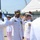 USS South Dakota (SSN 790) Holds Change of Command Ceremony A formal U.S. Navy military ceremony is taking place at a naval installation, likely a pier or shipyard. Multiple naval personnel in white dress uniforms are exchanging salutes, with a senior officer — identifiable by gold insignia, command device on cap, and multiple service medals — at the center. The setting includes industrial port infrastructure and a white event tent in the background.