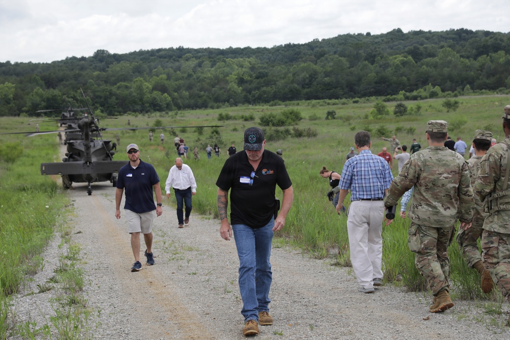 A group of civilians and military personnel in camouflage uniforms walk along a gravel path in a rural, grassy field setting. A military helicopter, appearing to be an AH-64 Apache, is visible on the left side of the image. The scene suggests an organized military demonstration, base tour, or civilian-military engagement event.