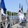 USS South Dakota (SSN 790) Holds Change of Command Ceremony A U.S. Navy color guard in dress white uniforms marches across a gangway or pier ramp, carrying the American flag and what appears to be a unit or command flag. The ceremony appears to be taking place at a naval installation near a body of water, with wooded hills visible in the background. The scene is consistent with a formal military commissioning, decommissioning, or change of command ceremony.