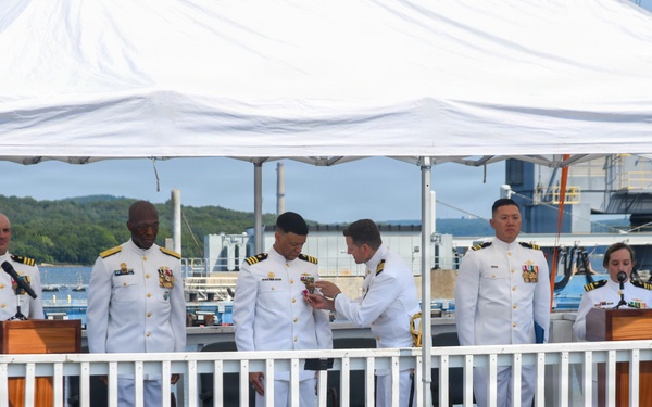A formal U.S. Navy ceremony is taking place on a platform labeled 'USS South Dakota,' with several naval officers in dress white uniforms gathered under a white canopy tent. The event appears to be a commissioning, change of command, or award ceremony conducted at a waterfront naval facility. Two officers stand at wooden podiums while others are engaged in what appears to be a pinning or award presentation in the center.
