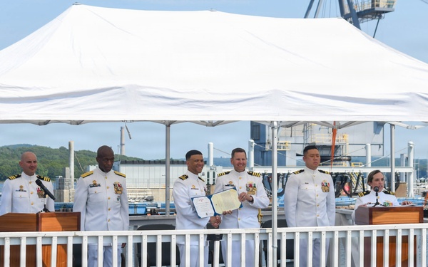A formal U.S. Navy ceremony is taking place on a platform bearing the name 'USS South Dakota.' Five naval officers in white dress uniforms are present, with two individuals at the center appearing to exchange or present an official document or certificate. The event is held outdoors at a naval facility with water and industrial port infrastructure visible in the background.