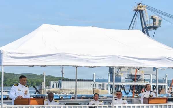 A formal U.S. Navy ceremony is taking place aboard or alongside the USS South Dakota submarine, identifiable by the nameplate visible on the platform. Several naval officers in white dress uniforms are seated on a raised ceremonial platform under a white canopy tent, with one officer speaking at a podium. The setting appears to be a naval shipyard or submarine base, with industrial cranes and waterfront facilities visible in the background.