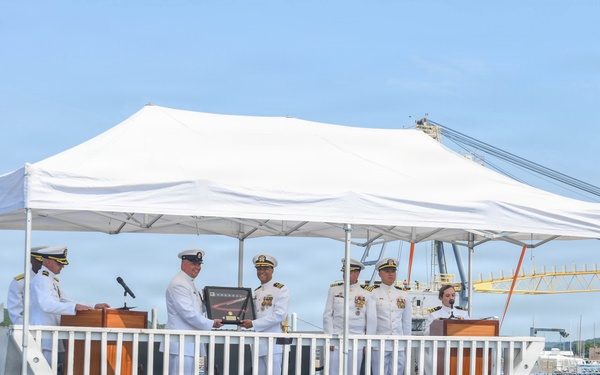 A formal U.S. Navy ceremony is taking place on the deck of the submarine USS South Dakota, as identified by the orange nameplate visible on the platform. Several naval officers in white dress uniforms are gathered under a white canopy tent, with two individuals appearing to exchange or present an award or plaque. The event appears to be a commissioning, change of command, or similar official military ceremony conducted at a naval facility.