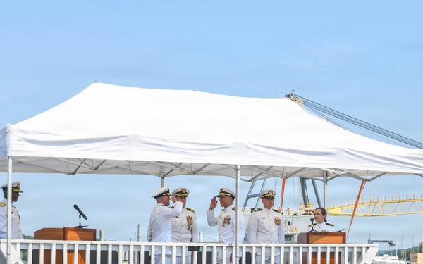 A formal U.S. Navy commissioning or change-of-command ceremony is taking place aboard the USS South Dakota submarine. Several naval officers in white dress uniforms are on a decorated platform, with some rendering salutes. The event appears to be conducted at a naval base with waterfront facilities visible in the background.