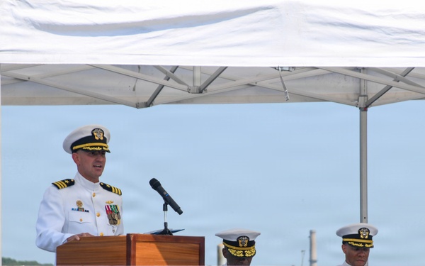 A U.S. Navy officer in dress white uniform is delivering a speech at a podium during what appears to be a formal military ceremony. Three other naval officers in dress whites are seated in the background under a white canopy tent. The setting appears to be an outdoor waterfront location.