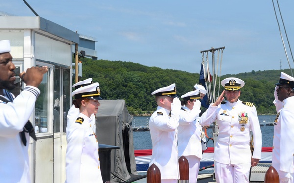 A formal U.S. Navy ceremony is taking place on a pier or dock, featuring personnel in white dress uniforms rendering salutes along a red carpet. The scene appears to be a change of command, commissioning, or official arrival/departure ceremony. Multiple officers and enlisted sailors are positioned in a ceremonial formation near the waterfront.